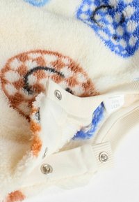 Weiche, cremefarbene Babykleidung mit Druckknöpfen und felligen braunen und blauen Kreismustern.