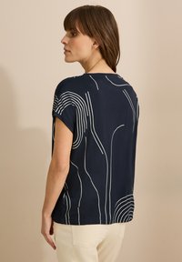 Námořnická modrá blůza s krátkým rukávem se bílými abstraktními liniemi. Měkký materiál s volným střihem a bočními rozparky na spodním lemu.