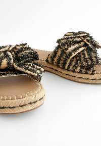 Sandales beige tissées à enfiler avec des rayures texturées noires et de grands nœuds décoratifs sur le dessus, avec des semelles entourées de corde et des bords cousus.