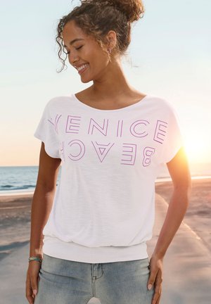 Weißes T-Shirt mit kurzen Ärmeln und dem Text "VENICE BEACH" in fetter lila Schrift. Es hat einen lässigen Schnitt und einen gerafften Saum.