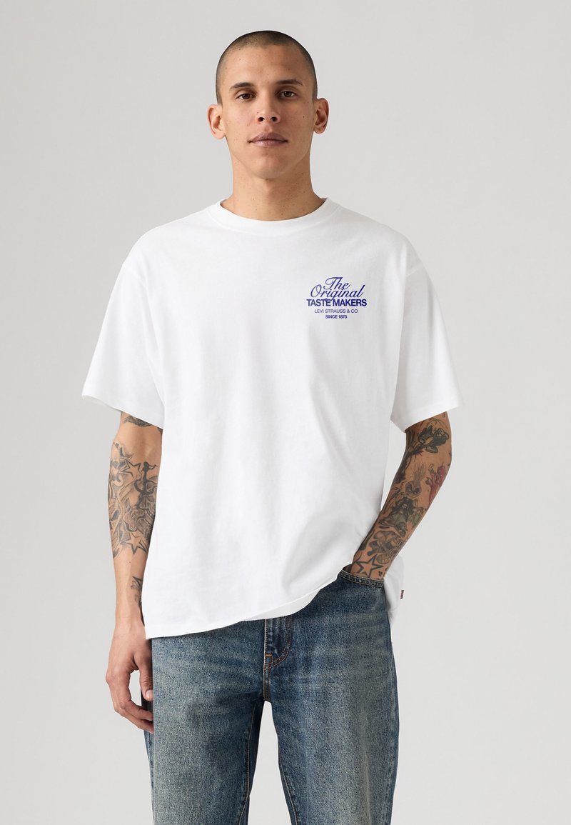 Giovane uomo con tatuaggi che indossa una maglietta bianca con testo blu "The Original Taste Makers Levi Strauss & Co Since 1873" e jeans blu.