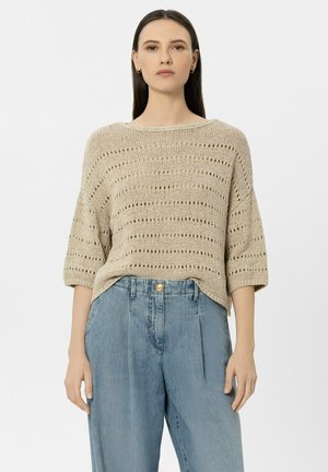 Woman with straight dark hair wearing a beige open-knit sweater and high-waisted relaxed-fit blue jeans standing against white background.