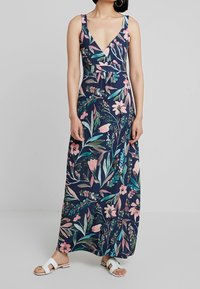 Kobieta w bezrękawnej niebieskiej sukience maxi w różowy i zielony kwiatowy wzór oraz białych płaskich sandałach na otwartym palcu, stojąca na tle jednolitym.