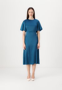 Robe en satin turquoise avec des manches courtes et légères, un corsage ajusté et une jupe midi. Texture lisse, design minimal, accompagnée de chaussures à talons de couleur claire.