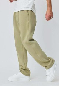 Pantalon de survêtement vert olive clair avec une texture douce, design fuselé, deux poches latérales et taille à cordon, associé à des baskets blanches.