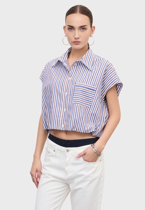 Žena oblečená v krátkej modro-hnedej pruhovanej košeli s krátkym rukávom a bielymi nohavicami, s kruhovými náušnicami a strieborným náramkom.