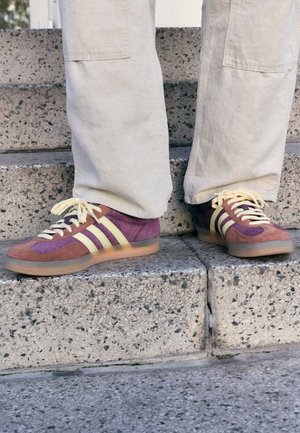 Person iført beige bukser og lilla og brune sneakers med gule striber, der står på betonstrappe.
