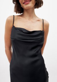 Nainen, joka on pukeutunut mustaan satiiniseen liukuvaatteeseen, jossa on ohuet olkaimet ja laskostettu kaula-aukko, hymyilee lempeästi yksinkertaista valkoista taustaa vasten.