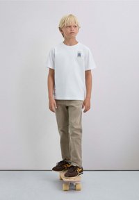 Witte katoenen t-shirt met een blauw en geel grafisch ontwerp, beige broek, staand op een skateboard. Bruine schoenen met gele accenten.