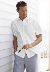 Camicia bianca a maniche corte con bottone e una tasca sul petto. Chi la indossa tiene degli occhiali da sole e si abbina a pantaloni grigio chiaro.
