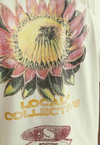 Šviesiai kremas audinys su dideliu gėlių raštu, turinčiu geltoną centrą, rožinius žiedlapius ir užrašą "LOCAL* COLLECTIVE" auksine spalva.