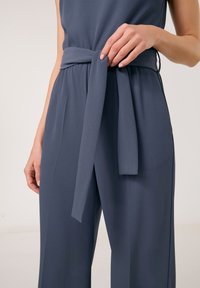 Dunkelgrauer Overall mit glatter Textur, ausgestattet mit einem gebundenen Gürtel in der Taille und weit geschnittenen Beinen. Klare Linien und taillierte Passform.