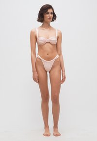 Γυναίκα όρθια, ξυπόλυτη, που φορά σετ μπικίνι σε ανοιχτό ροζ με ρυτιδωτή υφή και ψηλά κομμένα σλιπ, με φόντο απλό λευκό.