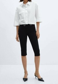 Camicetta bianca corta con bottoni e tasca frontale, abbinata a pantaloni capri neri aderenti e décolleté nere con punta affusolata.