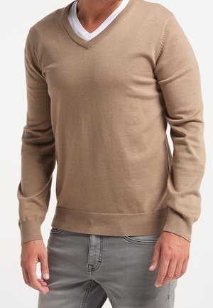 Mężczyzna ubrany w beżowy sweter z dekoltem w serek nałożony na białą koszulkę, w połączeniu z szarymi dżinsami na jednolitym białym tle.