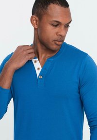 Mies, który nosi niebieską koszulę henley z długim rękawem i brązowymi guzikami, patrzy w dół, poprawiając kołnierzyk prawą ręką na jednolitym tle.