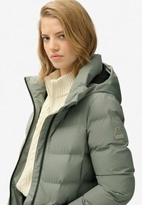 Dik, olijfgroene parka met ritssluiting en een capuchon. Is voorzien van een geribbeld crèmekleurig truitje eronder en een logopatch op de linkermouw.