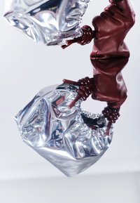 Petit sac argenté brillant avec des poignées à cordon en cuir marron reflétant sur une surface blanche contre un fond blanc.