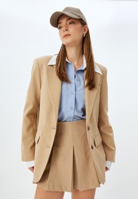 Beige blazer med to knapper, hvid krave, blå button-up skjorte og beige plisseret nederdel. Matchende ternet kasket fuldender sættet.