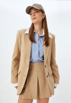 Blazer beige con due bottoni, colletto bianco, camicia blu con bottoni e gonna plissettata beige. Un berretto a scacchi coordinato completa l'insieme.