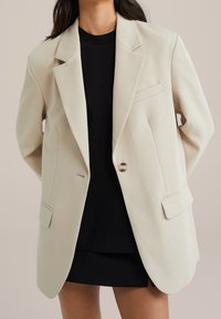 Beige skräddarsydd kavaj med enknappsstängning, slagkrage, två framfickor och en slät yta, bärs över en svart topp.