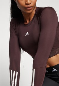 Smeđi sportski majica dugih rukava s bijelim Adidas logotipom na prsima. Ima bijele pruge na rukavima i rebraste detalje. Elastčan, prilagođen dizajn.