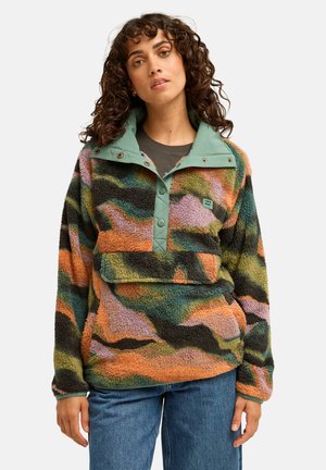 Femme aux cheveux bouclés portant un pull polaire camouflage multicolore avec un col vert et des pressions, associé à un jean bleu, sur un fond uni.