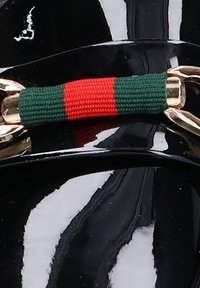 Material negro brillante con herrajes dorados, que presenta un detalle textil de rayas verdes y rojas. Superficie suave con un brillo sutil; acentos decorativos.