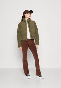 Mulher vestindo uma jaqueta acolchoada verde-oliva, blusa branca, calças castanhas, boné preto e tênis preto e branco, em pé contra um fundo branco liso.