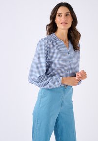 Blusa a scacchi blu e bianca con scollatura a V, chiusura con bottoni e maniche a palloncino, abbinata a jeans chiari a vita alta.