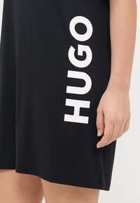 Juodas suknelė su vertikalia baltos spalvos "HUGO" logo kairėje pusėje. Medžiaga atrodo lygi, su laisvu siluetu ir standartiniu apvadu.