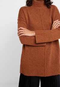 Maglione marrone lavorato a maglia con una scollatura alta, polsini a coste e una vestibilità morbida. Presenta un dettaglio a righe sulla parte anteriore e una texture morbida.
