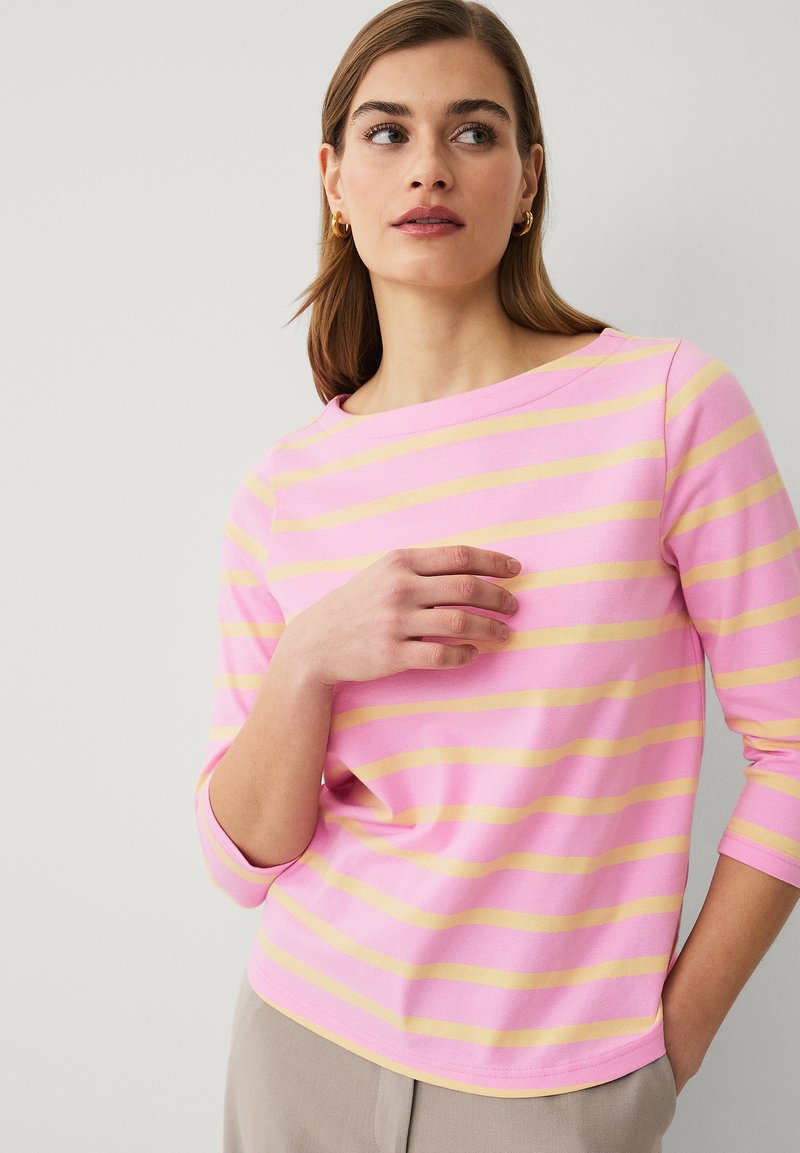 Kvinde med lyst brunt hår iført en pink og gul stribet langærmet trøje, hånd hvilende på brystet, ser til siden.