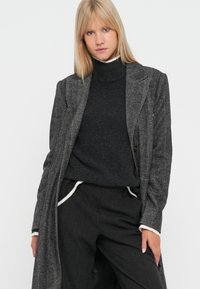 Cappotto grigio a scacchi con collo lungo, indossato sopra un maglione a collo alto scuro, abbinato a pantaloni scuri con dettagli chiari.