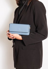 Clutch rettangolare blu chiaro con una texture liscia, caratterizzato da una chiusura a patta e dettagli in metallo dorato, tenuto tra le mani accanto a un blazer nero.