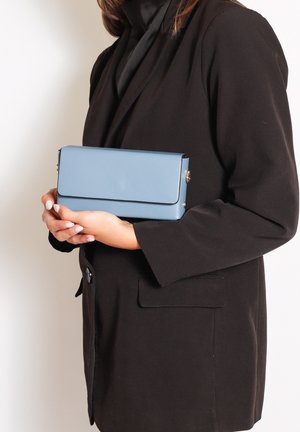 Hellblaues rechteckiges Clutch mit glatter Textur, das über einen Klappenverschluss und goldene Hardware-Details verfügt, in den Händen gehalten vor einem schwarzen Blazer.