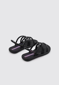 Chanclas negras con múltiples tiras delgadas sobre una plantilla texturizada. Presentan una etiqueta morada en la suela con el nombre de la marca. Material de goma.