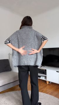 Didelis pilkas megztas liemenė su aukštu apykakliu, raštuotos tekstūros ir atviromis šoninėmis dalimis. Modelis dėvi dryžuotą mar škinius ir juodas kelnės.