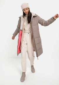 Gewatteerde lange jas in taupe met een roze voering, voorzien van een capuchon en een rits aan de voorkant. Gepaard met beige broeken en beige veterschoenen.