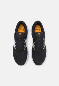 Chaussures de sport noires avec une tige en mesh, des accents jaunes et une semelle blanche. Elles comportent des lacets et un col rembourré pour un meilleur maintien.