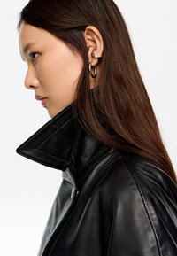 Melna ādas jaka ar augstu apkakli, ar gludu tekstūru un sudraba rāvējslēdzēja detaļām. Modelim ir garas brūnas matu cirtas un sudraba riņķa auskari.