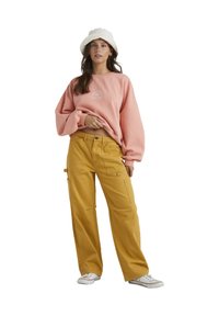Rosa sweatshirt med en rund logotyp, kort i midjan, tillsammans med gula vida cargo-byxor med sidofickor. Vit bucket hat och skor.