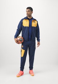 Marineblauw en gele sportieve trainingsoutfit met een ritssluitingjack en bijpassende broek. Inclusief een gestreept shirt, een sportbal en roze voetbalschoenen.