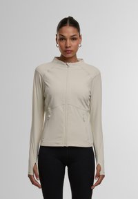 Lyse beige zip-up jakke med ståkrave, der har to lynlåslommer på siderne og lange ærmer med tommelhuller. Glat, strækbart stof.