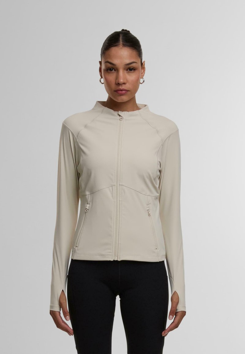 Lyse beige zip-up jakke med ståkrave, der har to lynlåslommer på siderne og lange ærmer med tommelhuller. Glat, strækbart stof.