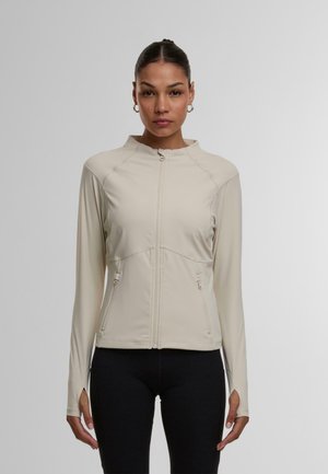 Světlobéžová bunda na zip s stojáčkem, která má dvě kapsy na zip po stranách a dlouhé rukávy s otvory na palce. Hladký, pružný materiál.