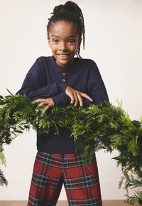 Navy long-sleeve shirt with three wooden buttons, paired with red plaid pants. Hands resting on green foliage garland. Neutral background.