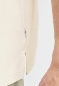 T-shirt en coton beige avec une fente sur le côté au niveau de l'ourlet et une petite étiquette sur le côté. Texture lisse et coupe droite.