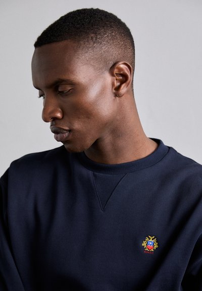 Navy blue sweatshirt with a round neckline and ribbed cuffs, featuring a colorful embroidered logo on the left chest. Smooth cotton fabric.