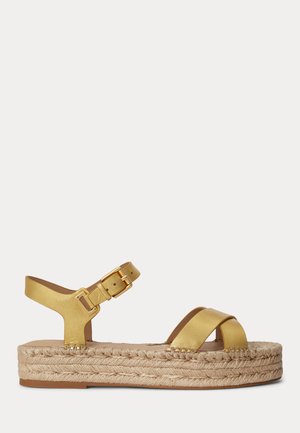 Sandalia de plataforma de cuero dorado con correa en el tobillo y hebilla, suela de yute trenzado y tiras cruzadas sobre los dedos, vista lateral sobre fondo blanco.